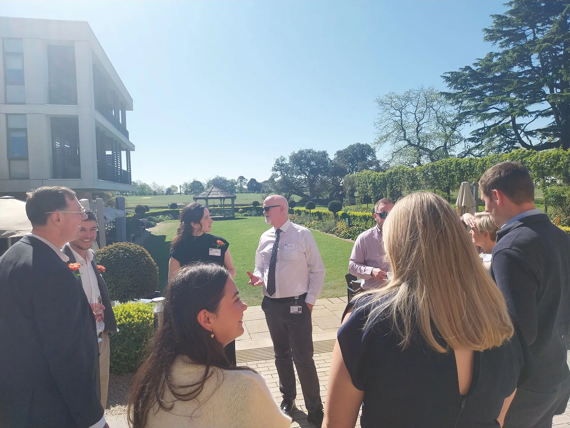 A group of people chatting, standing on a sunny terrace overlooking gardens